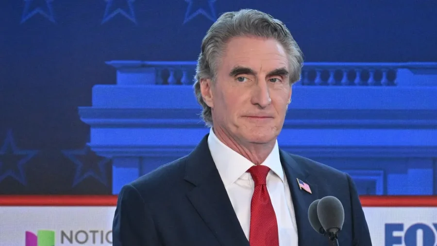 Un hombre de traje oscuro, corbata roja y un pin de la bandera de Estados Unidos, de pie frente a un micrófono en un set de televisión