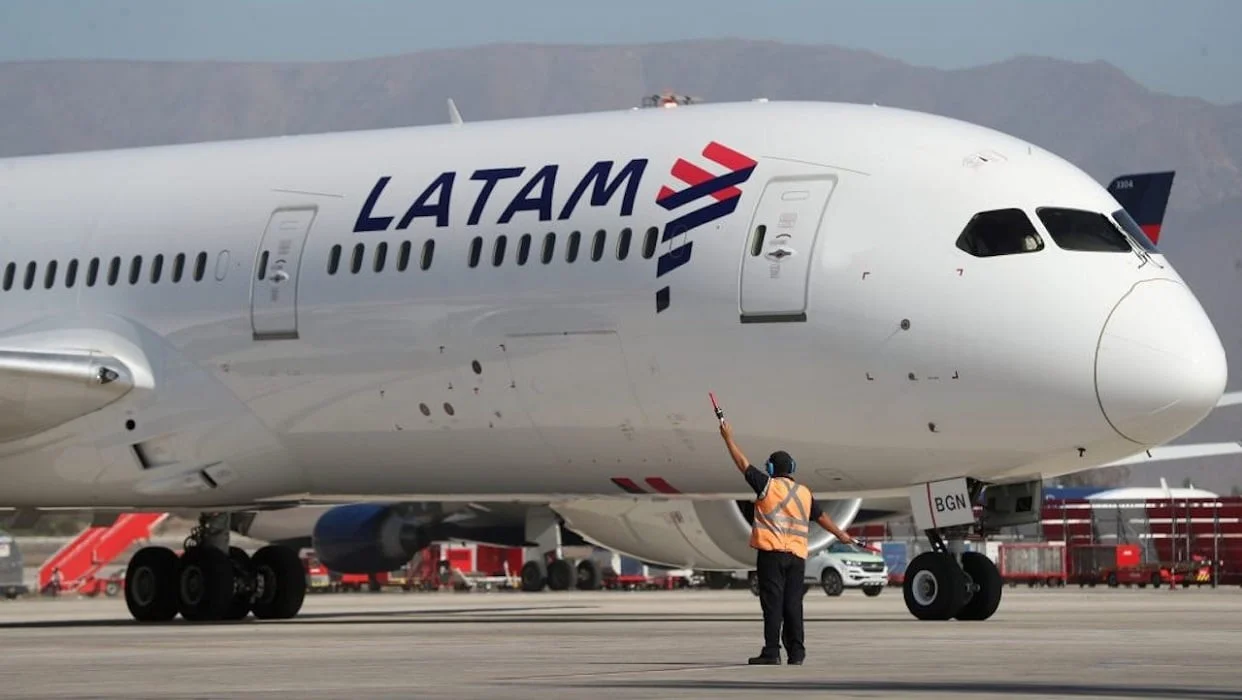 Un operario con chaleco naranja hace señales con una paleta frente a un avión de Latam en la plataforma de un aeropuerto