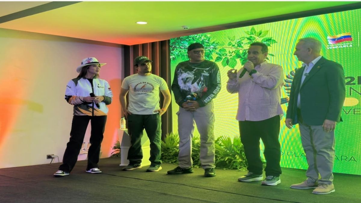 Cinco personas en una tarima frente a una pantalla verde con ilustraciones de hojas; uno de ellos habla con un micrófono
