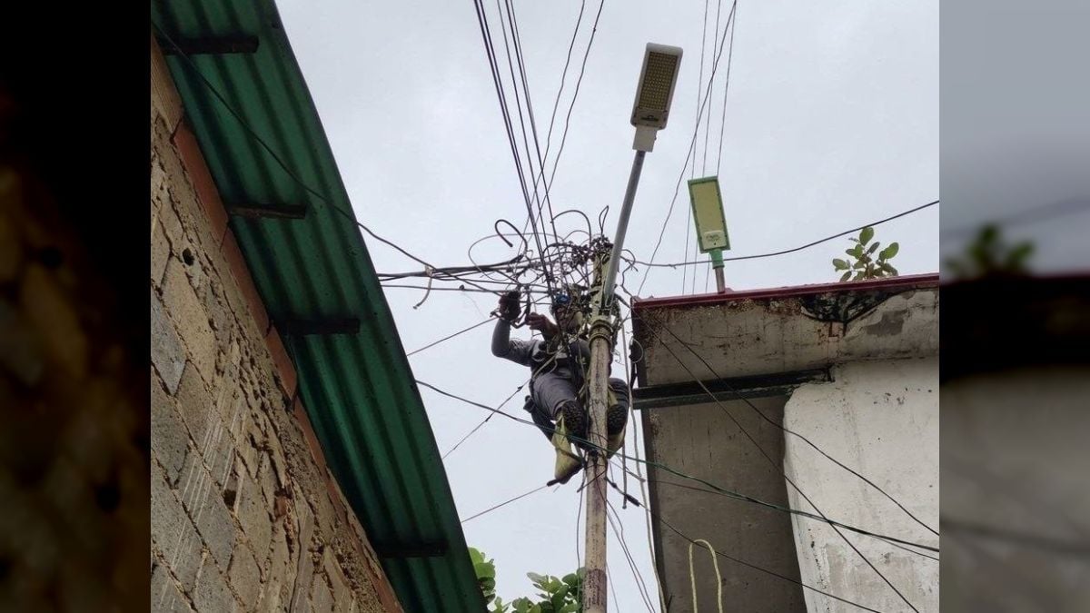 Un trabajador encaramado en un poste del tendido eléctrico, entre cables enredados y techos de casas