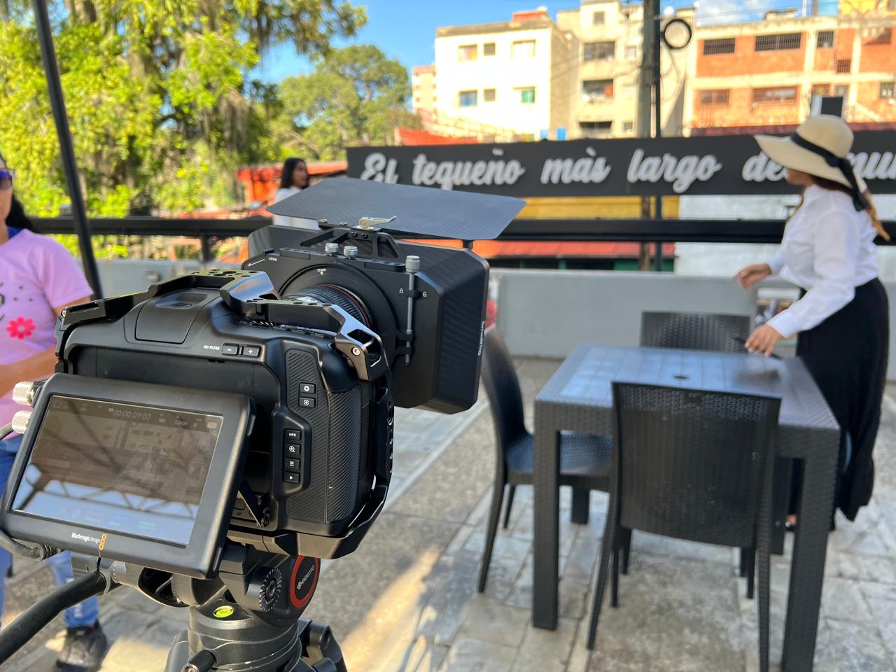 Una cámara de cine en primer plano durante un rodaje en una terraza, con un letrero que dice «El tequeño más largo» al fondo