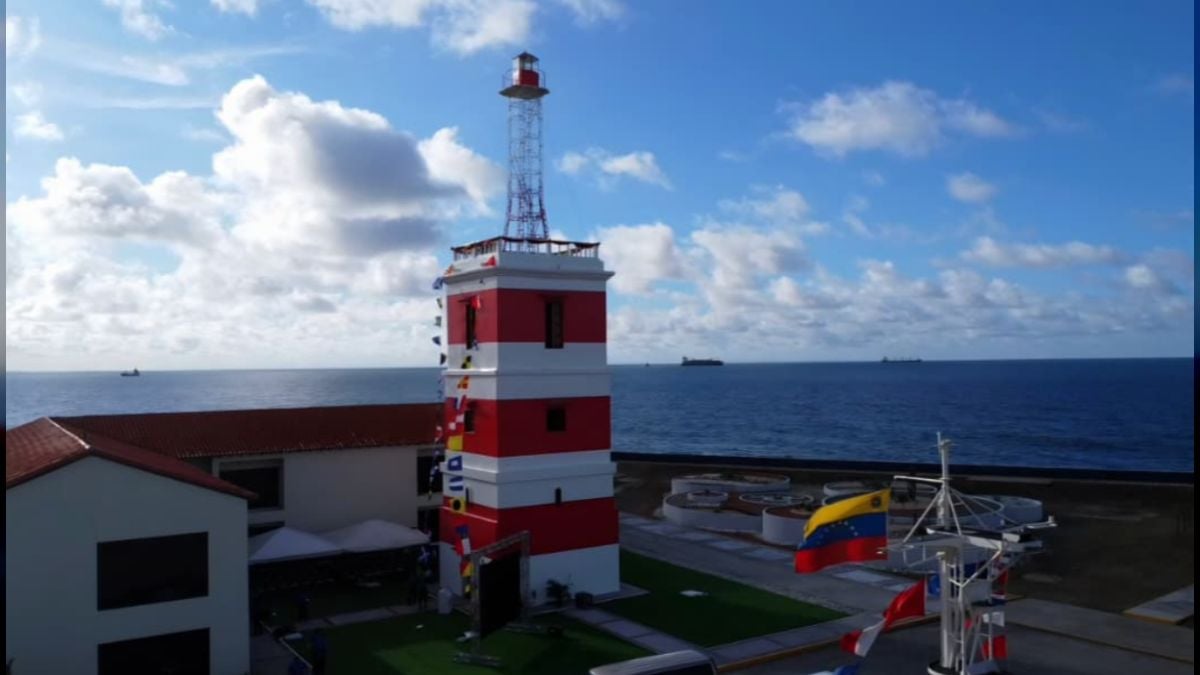 Un faro de franjas rojas y blancas, rematado por una torre de acero, junto a edificios de techo rojo frente al mar, con banderas de señales y una bandera de Venezuela en un mástil