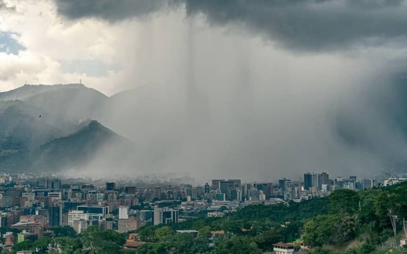 Vista panorámica de Caracas con edificios y cerros bajo un aguacero que cae desde nubes oscuras
