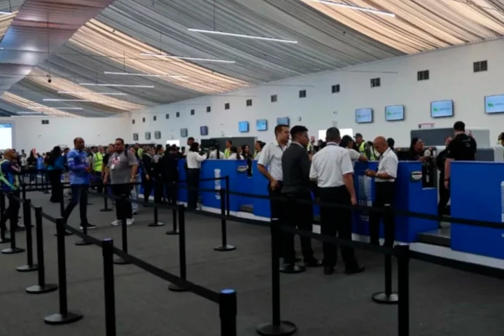 Pasajeros y trabajadores frente a los mostradores de una terminal con techo de lona, con cintas para hacer fila en primer plano