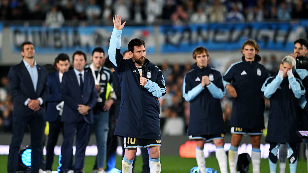 Un jugador de Argentina con el número 10 levanta la mano y habla con un micrófono sobre el césped de un estadio lleno, rodeado de compañeros y miembros del cuerpo técnico