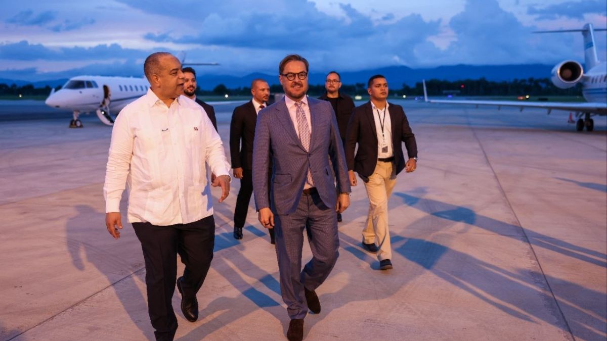 Un hombre de traje y lentes camina por la pista de un aeropuerto junto a un hombre de camisa blanca y una comitiva, con aviones privados al fondo