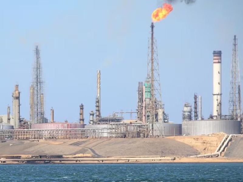 Instalaciones de una refinería a orillas del mar, con torres, chimeneas y tanques, y una llama encendida en lo alto de una torre