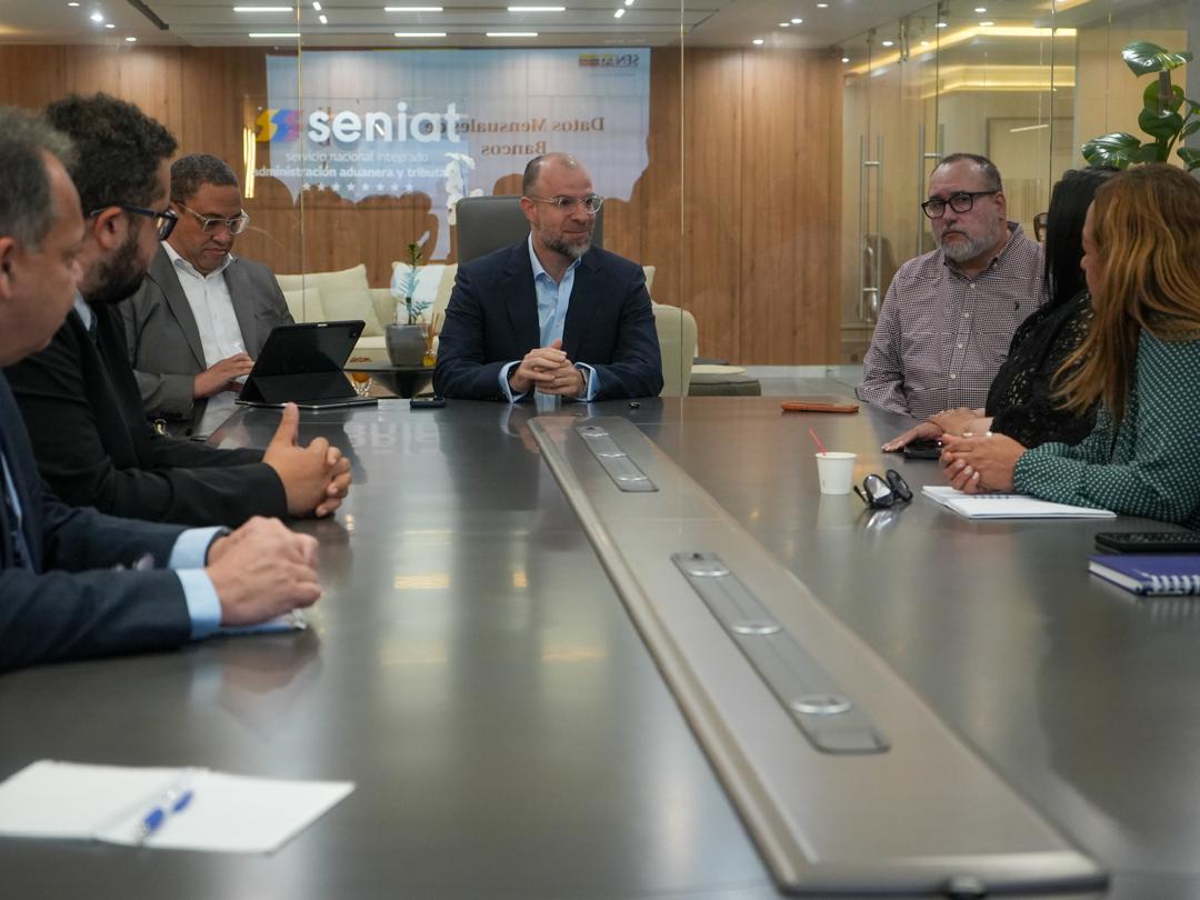 Varias personas reunidas alrededor de una mesa de juntas, con el logo del Seniat en una pantalla al fondo