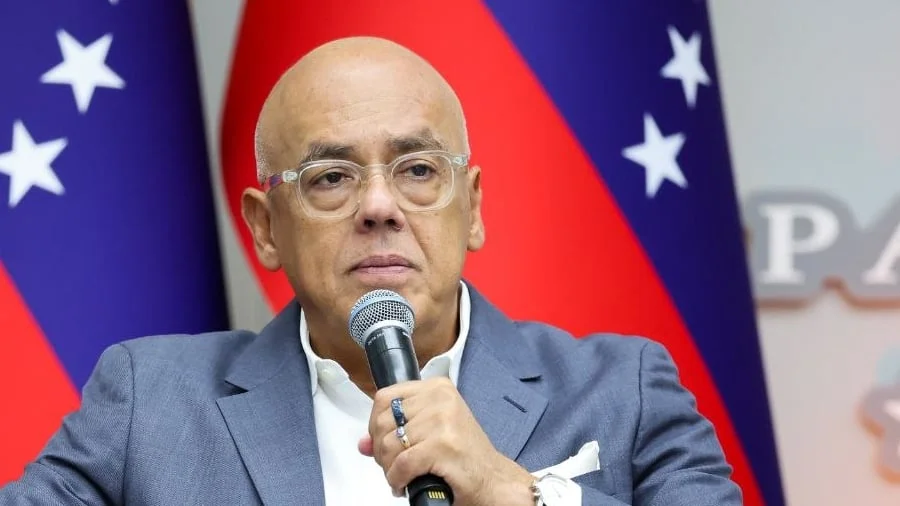 Un hombre con lentes y traje gris escucha con un micrófono en la mano, frente a banderas de Venezuela
