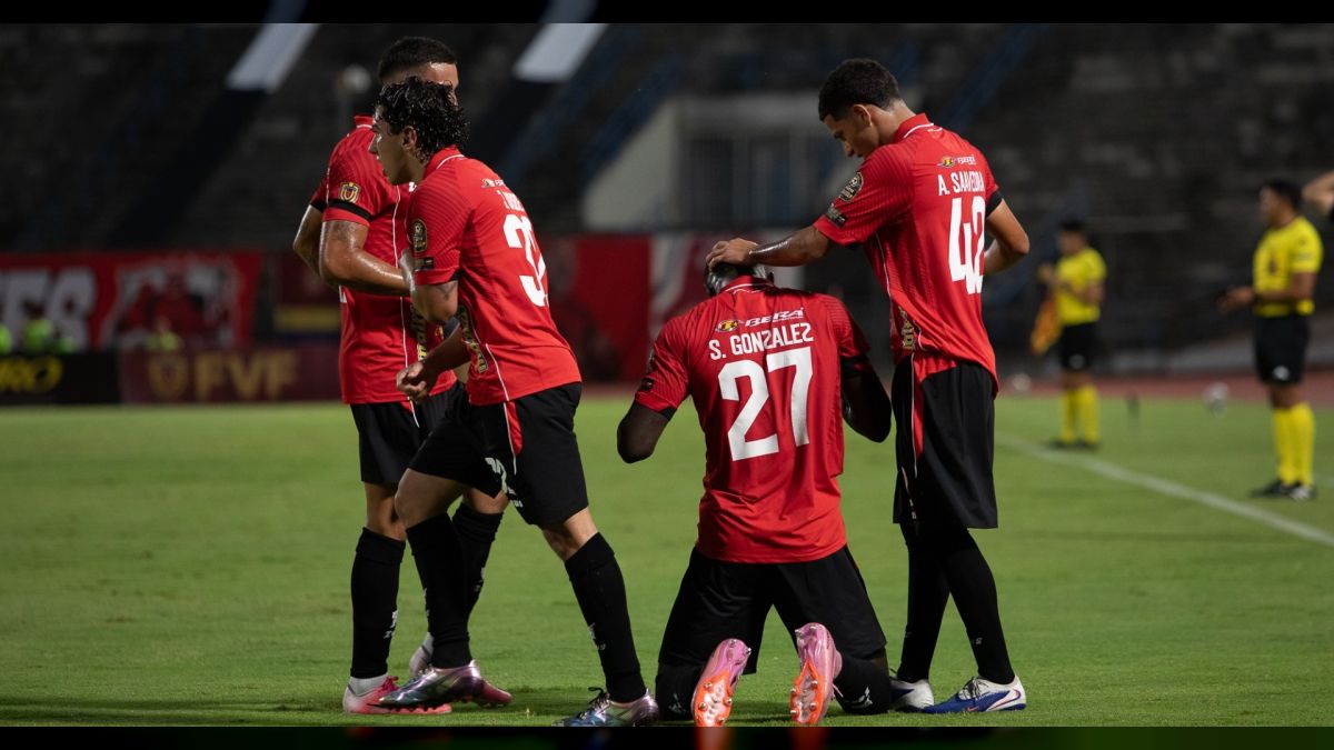 Jugadores de camiseta roja celebran en el césped de un estadio de noche; uno de ellos, con el número 27 y el nombre González en la espalda, está de rodillas mientras un compañero le pone la mano en la cabeza