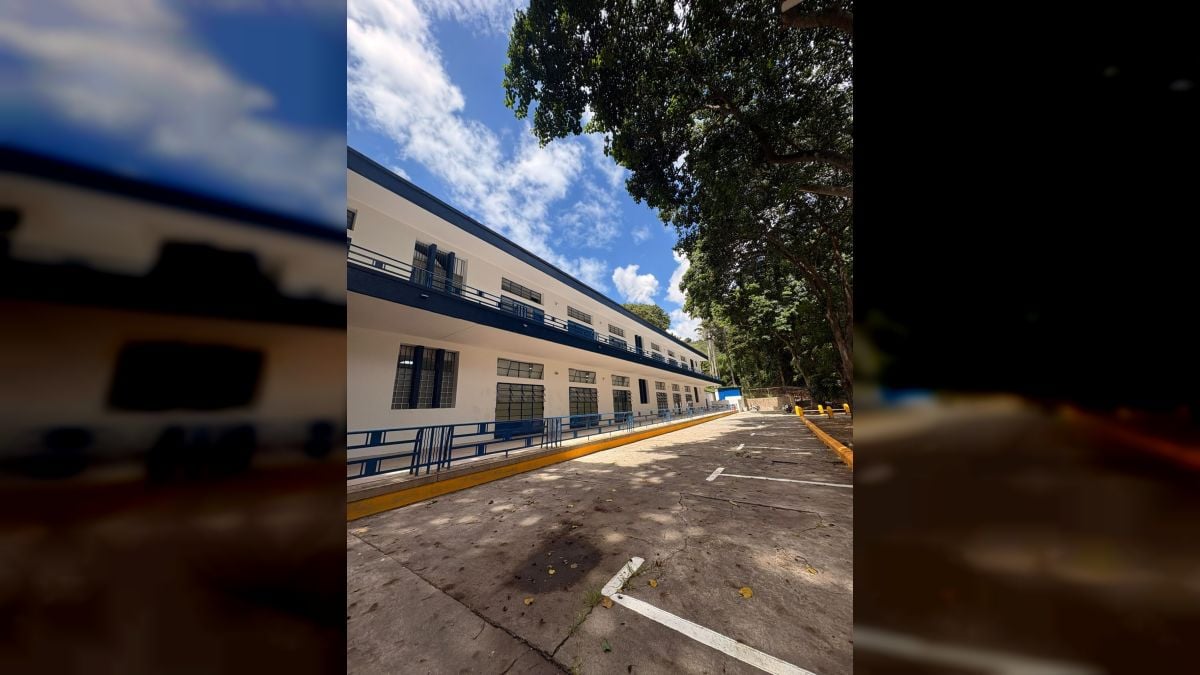 Un edificio blanco de dos plantas con barandas azules y ventanas con rejas, junto a un estacionamiento vacío y árboles