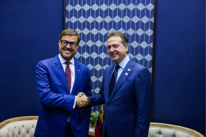Dos hombres de traje azul se dan la mano y sonríen frente a un panel con motivos geométricos, junto a una pequeña bandera de Venezuela