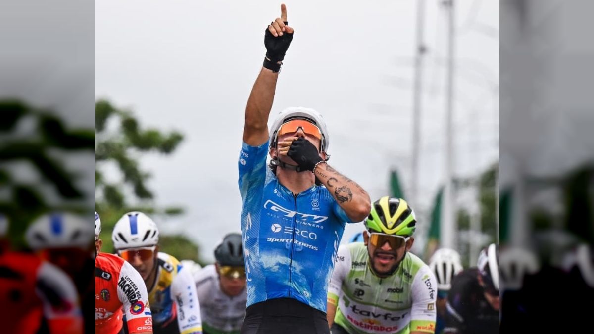 Un ciclista con maillot azul y casco blanco levanta el índice hacia el cielo y se lleva la otra mano a la boca, delante de otros corredores