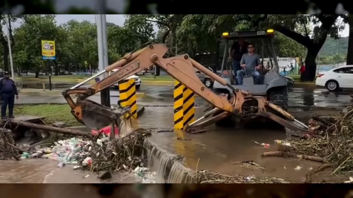 Una retroexcavadora retira ramas y basura acumuladas junto a un canal desbordado en una avenida mojada