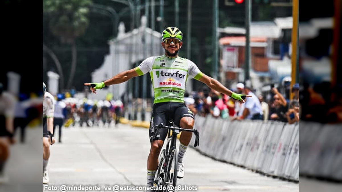 Un ciclista con maillot blanco y verde y casco verde abre los brazos sobre la bicicleta en una calle, con el público detrás de las vallas