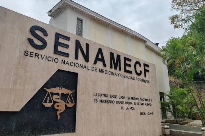 Fachada del Servicio Nacional de Medicina y Ciencias Forenses, con el nombre en letras grandes sobre un muro claro, un emblema dorado con una balanza y vegetación a la derecha