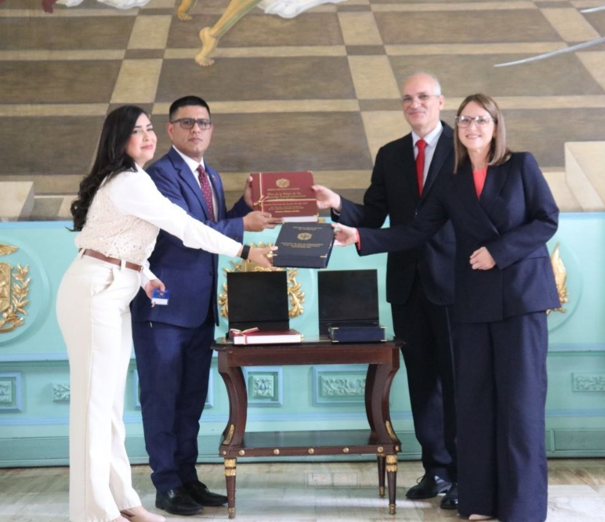 Dos mujeres y dos hombres de traje sostienen los tomos encuadernados del proyecto de presupuesto junto a una mesa con más documentos, en un salón con un mural en el techo
