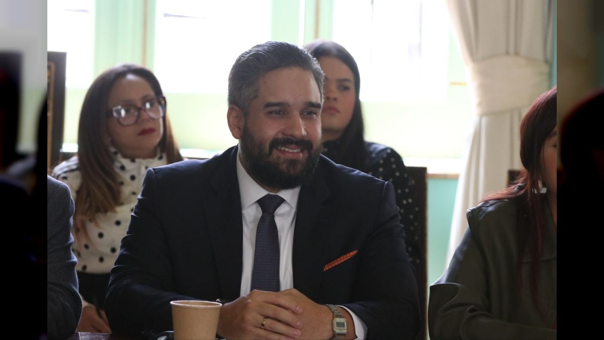 Un hombre de traje y corbata sonríe sentado a una mesa, con un vaso de papel delante y varias mujeres sentadas detrás