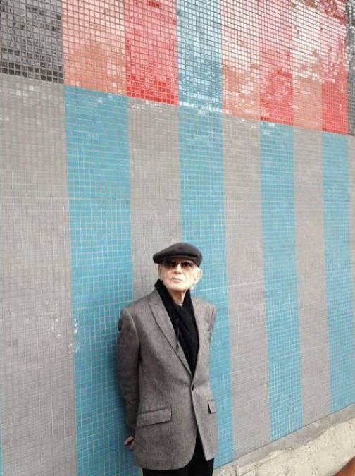 Mateo Manaure, con boina y chaqueta gris, frente a un mural de mosaicos