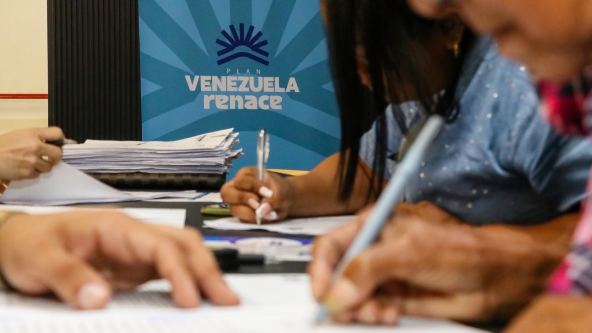 Personas firman documentos en una mesa frente a un pendón del Plan Venezuela Renace