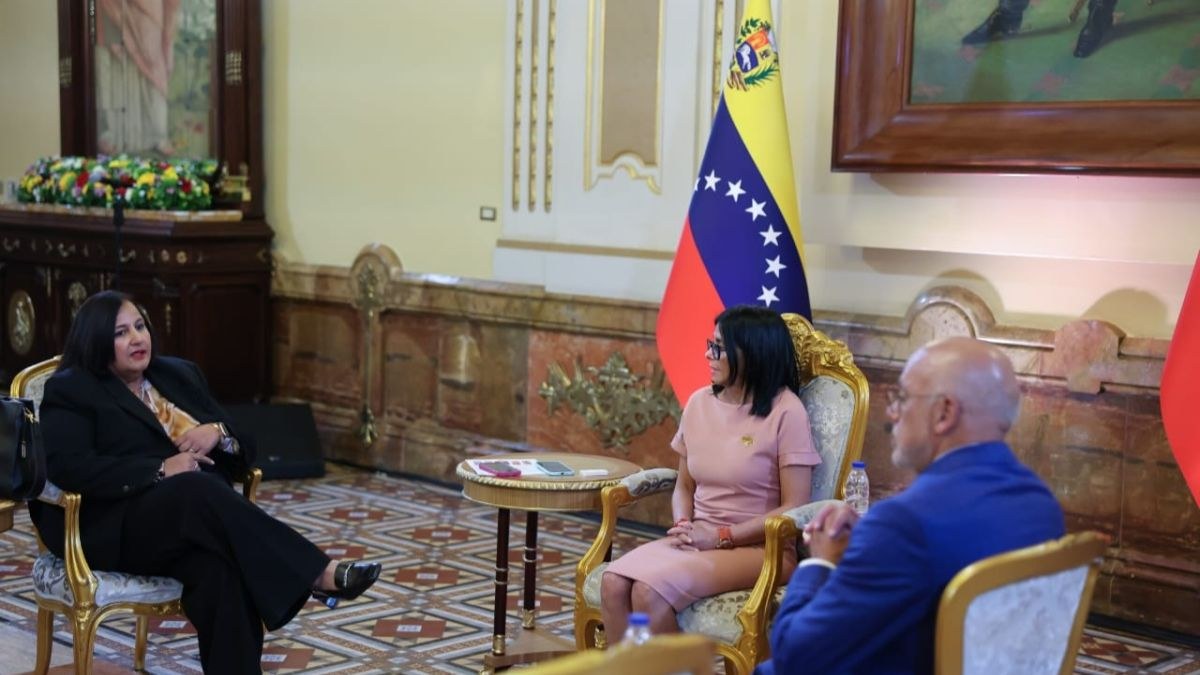 Tres personas conversan sentadas en sillones dorados en un salón, junto a una bandera de Venezuela