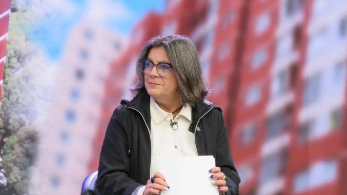 La ministra Paola Posani, con lentes y chaqueta negra, sostiene unas hojas frente a una imagen de edificios