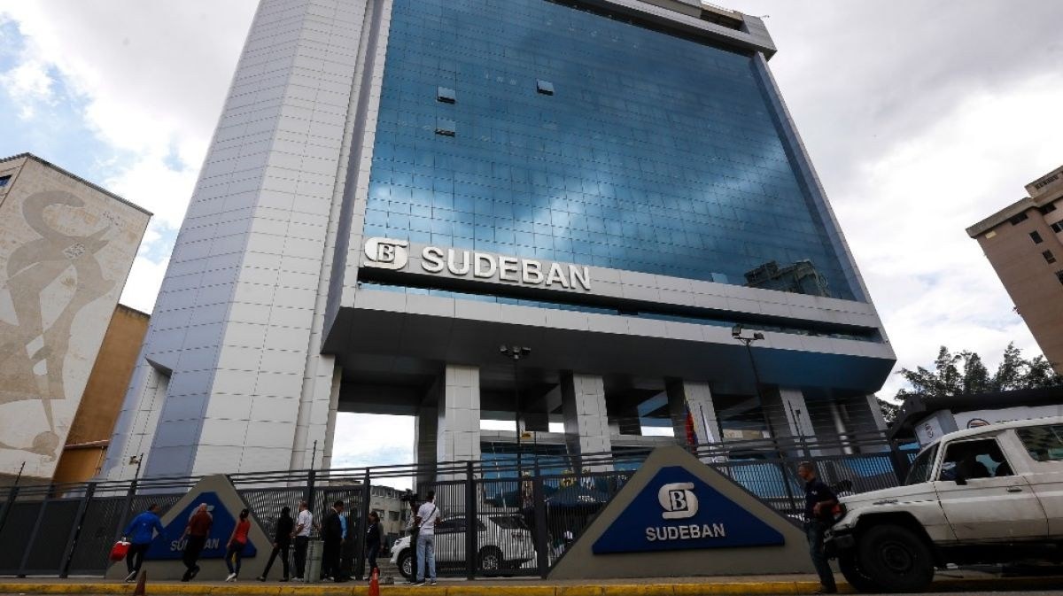 Edificio de fachada de vidrio con el letrero de Sudeban; varias personas caminan frente a la reja
