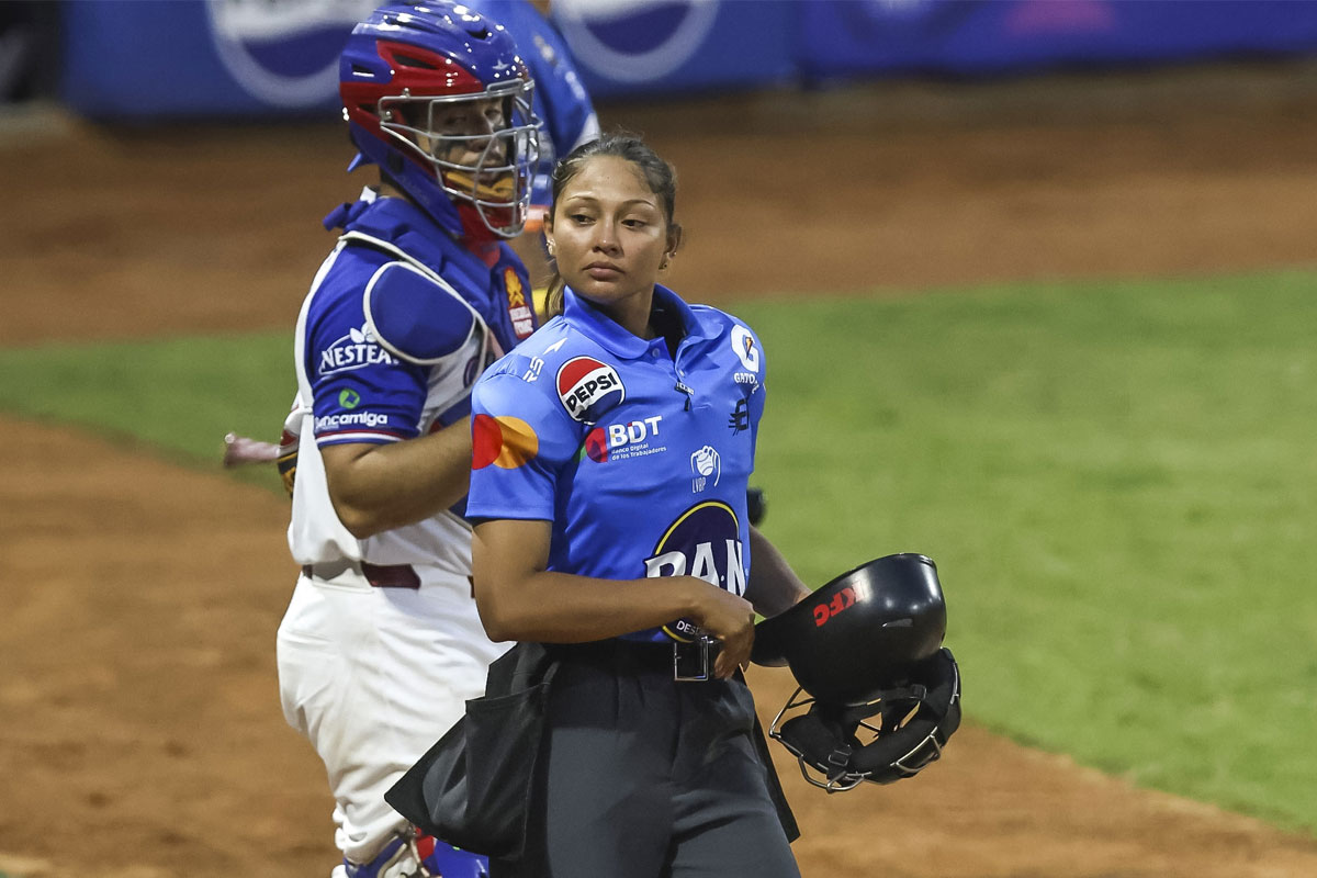 Una umpire con el casco en la mano camina en el terreno junto a un receptor