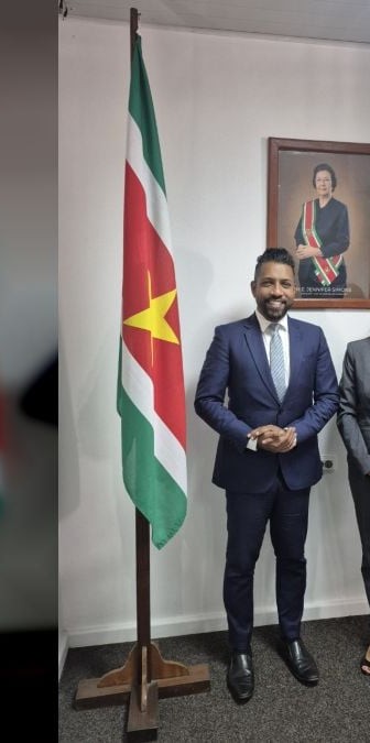 Un hombre y una mujer posan de pie junto a una bandera de Suriname en una oficina