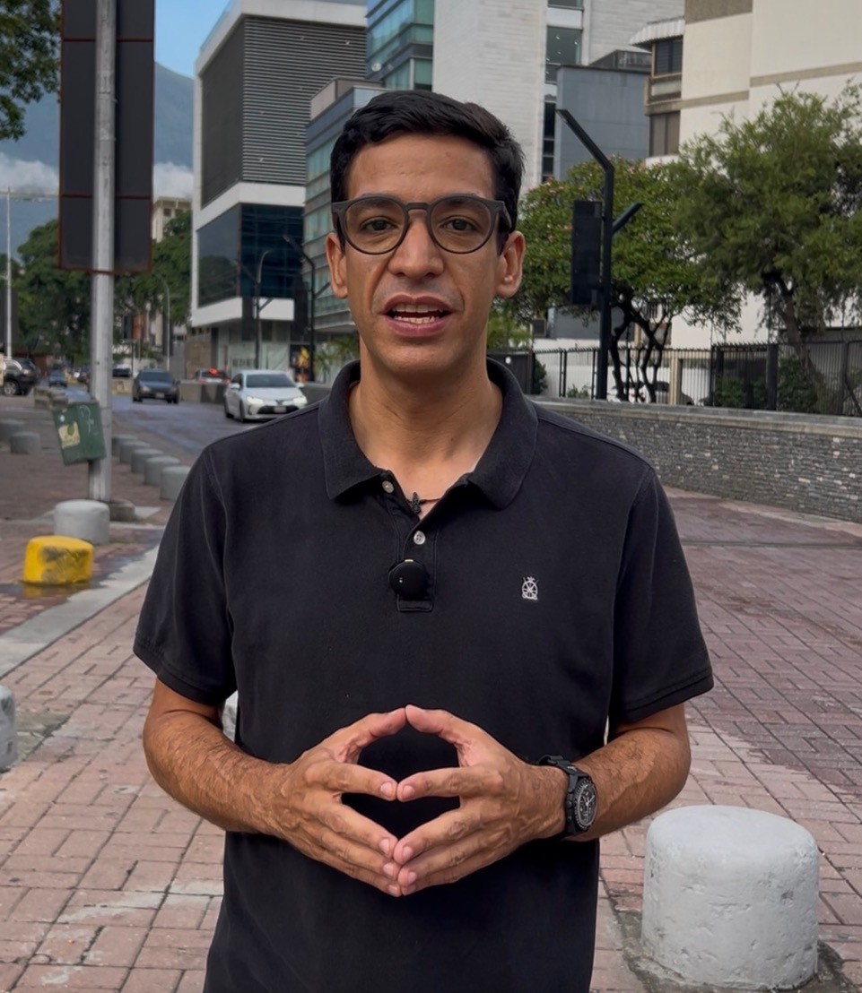 Un hombre con lentes y camisa tipo polo negra habla en una acera junto a una avenida con edificios de oficinas