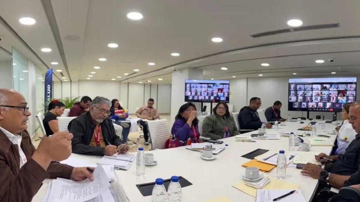 Funcionarios reunidos alrededor de una mesa, con dos pantallas de videoconferencia al fondo